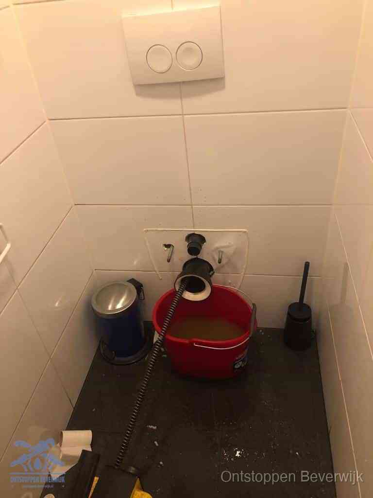 Een ontstopte wand-wc met verwijderde toiletpot waar een ontstoppingsveer in de wanduitgang verdwijnt, het achterhalen waar de verstopping zit in Beverwijk