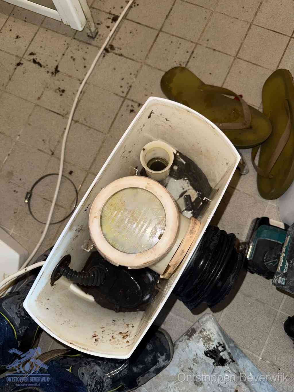 Een gedemonteerde toiletpot op de vloer met ontstoppingsveer en gereedschap ernaast, het verwijderen van een vastzittend voorwerp uit de wc in Beverwijk