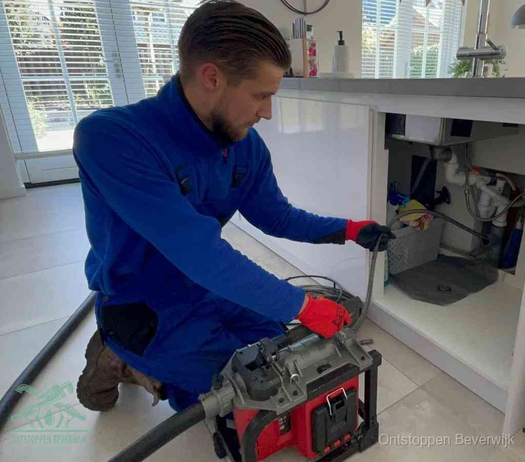 Een monteur met rode handschoenen knielt in een keuken en voert een elektrische ontstoppingsveer in tijdens het professioneel afvoer ontstoppen in Beverwijk